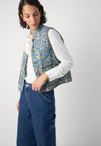 Γιλέκο με λουλουδάτο σχέδιο σε μπλε, χρυσό και πράσινο, χωρίς γιακά και με κουμπί, φορεμένο πάνω από λευκό τοπ και συνδυασμένο με τζιν παντελόνι.