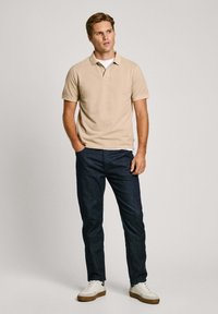 Bež polo majica s kratkim rukavima, u kombinaciji s tamnoplavim trapericama. Model nosi bijele i sive tenisice s teksturiranim đonom.