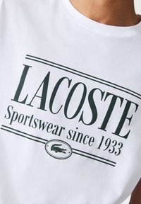 Bílý bavlněný tričko s zeleným nápisem "LACOSTE" a "Sportswear since 1933," s logem krokodýla v kruhovém designu.