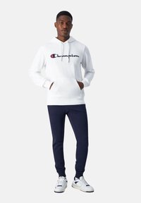 Witte katoenen hoodie met een voorzak en een Champion-logo, gecombineerd met navy joggingbroek en witte sneakers met navy accenten.