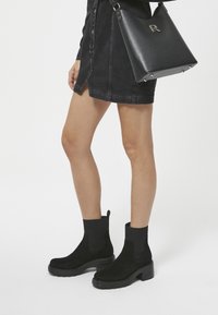Black suede ankle boots with ribbed sock collars and chunky rubber soles, paired with a black handbag featuring a logo detail.