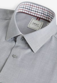 Camisa gris con un patrón, con un collar estructurado, que presenta un forro interior a cuadros en blanco y rojo, cierre de botones y una etiqueta.