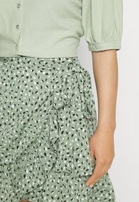 Blusa verde chiaro con maniche a sbuffo corte, abbinata a una gonna volant con un motivo astratto in bianco, nero e verde e un dettaglio a fiocco.