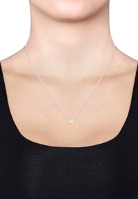 Sølv kæde med en pendants af stort bogstav "N", præsenteret mod en mørk tøjbaggrund. Enkelt, rent design.