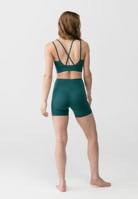 Naine kannab tehasinist ristsuunaliste rihmadega spordirinnahoidjat ja sobivaid kõrge vöökohaga rattapükse, seisab paljajalu ja on tagumikuga valge tausta poole.