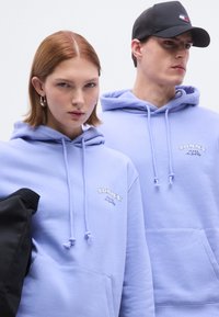 Hellviolette Kapuzenpullover mit Kordelzug und einer Fronttasche tragen das Logo "TOMMY JEANS". Eine Person trägt eine schwarze Kappe; beide haben eine glatte Textur.