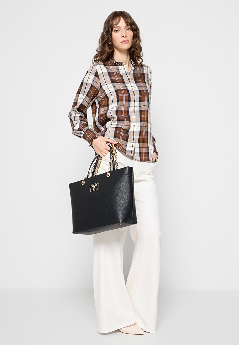 Sac en cuir noir avec des ferrures en ton doré et des accents en chaîne, associé à une chemise à carreaux bruns et blancs à boutons et à un pantalon blanc.