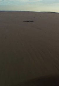 Et flatt, utstrakt sandlandskap under en klar blå himmel, med subtile teksturvariasjoner og en svak mørk flekk i forgrunnen.