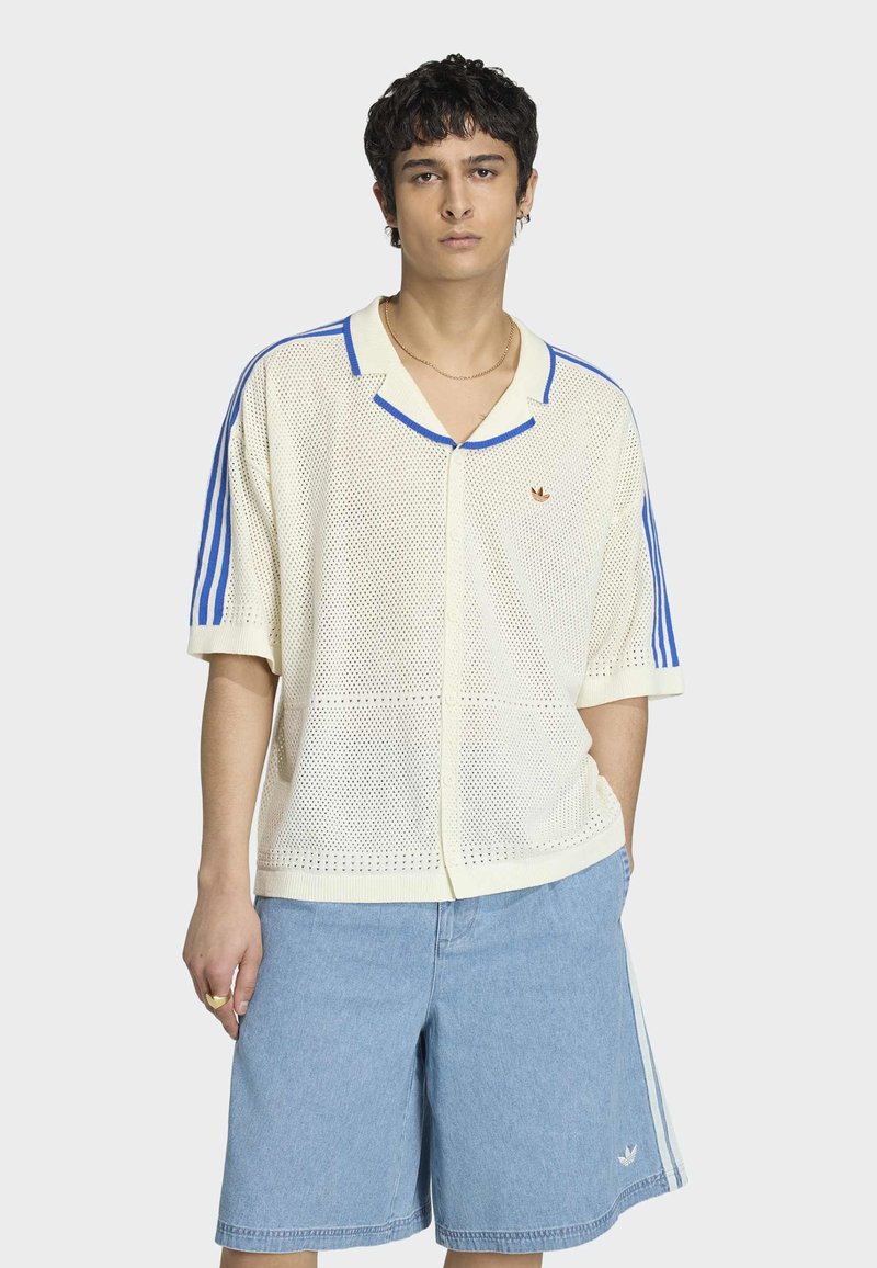 Jonge man die een crèmekleurige mesh korte mouw cardigan draagt met blauwe strepen en lichtblauwe spijkershorts met witte zijstrepen.