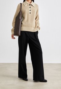 Vrouw in een beige gebreide trui en zwarte wijde broek, met een hoge donkerbruine leren tote tas over schouder.