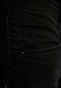 Jeans de mezclilla negra con una textura desgastada, sutiles detalles de costura y un pequeño bolsillo con herrajes de metal.