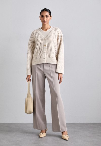Cardigan bej tricotat cu design cu țesătură deschisă, patru nasturi, asortat cu pantaloni largi gri deschis și balerini bej cu vârf ascuțit.