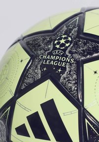 Nogometna lopta s neonsko zelenom površinom i crnim uzorcima zvijezda. Prikazuje logotip UEFA Lige prvaka i Adidas brendiranje u crnoj boji.