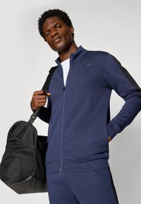 Casaco zip-up azul-marinho com detalhes em preto nos ombros, apresentando uma gola alta e um detalhe com o logotipo. O modelo segura uma bolsa de ginásio preta com rede.