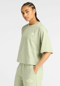 Mujer con el cabello trenzado, vistiendo una camiseta oversize de New Balance en verde claro y pantalones de chándal a juego con las manos en los bolsillos.