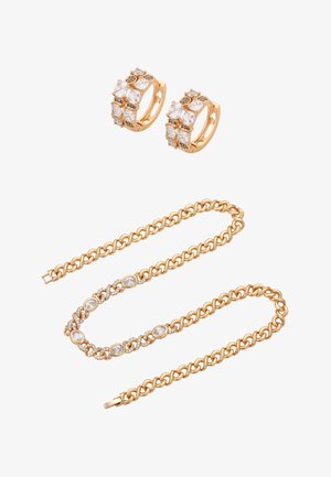 Para złotych kolczyków kołowych z przezroczystymi kamieniami, nad złotym naszyjnikiem łańcuszkowym z owalnymi i okrągłymi przezroczystymi kamieniami na białym tle.