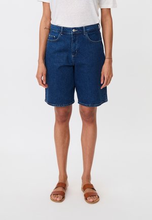 Persona con pantalones cortos de mezclilla azul oscuro hasta la rodilla, camiseta blanca y sandalias planas marrones, de pie frente a un fondo blanco liso.