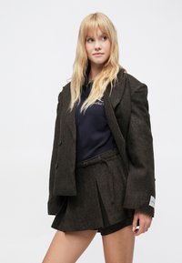 Barna tweed blézer nagy felgombolható gallérral és két gombbal, tengerészkék polóval és hozzáillő barna pliszírozott szoknyával.