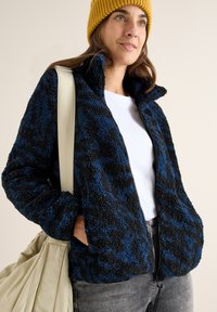Giacca in pile con zip, nera e blu, caratterizzata da un motivo testurizzato. Presenta un colletto alto e tasche laterali, abbinata a una camicia bianca.