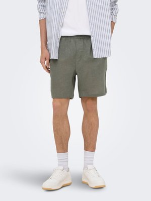 Homme portant un short vert olive, des baskets blanches à semelles en gomme, des chaussettes blanches, un t-shirt blanc et une chemise rayée bleue et blanche.