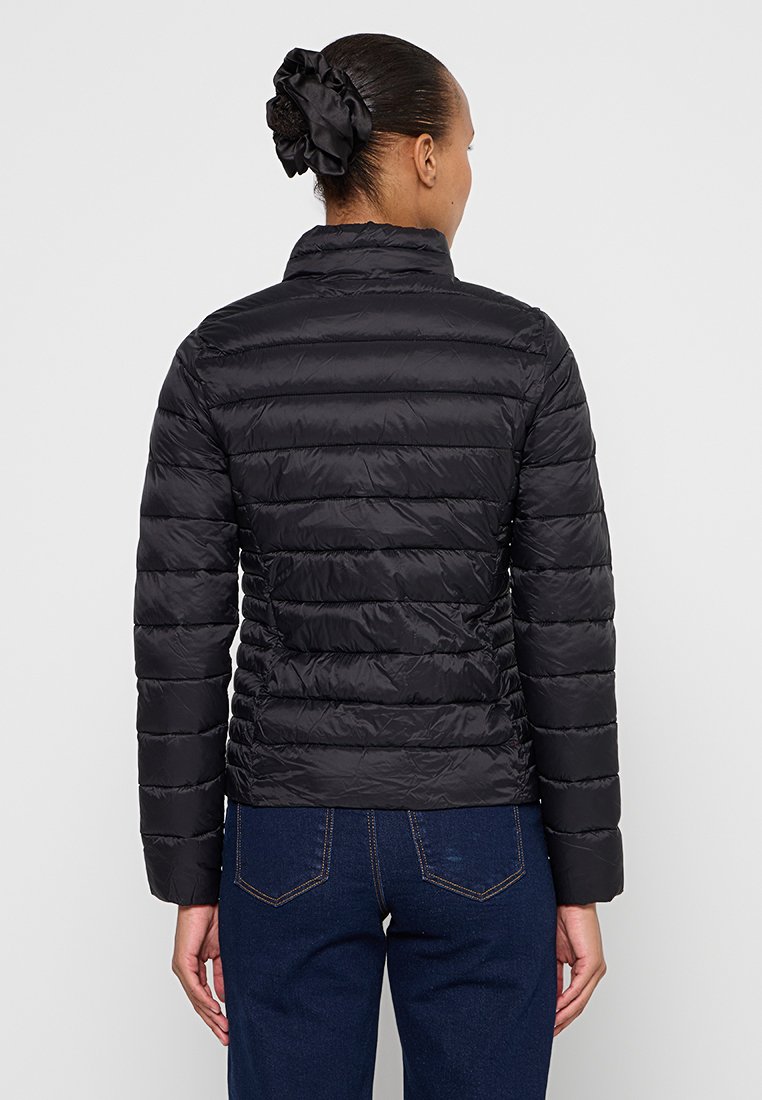 Schwarze isolierte Pufferjacke mit gerippter Textur, vertikalen Nähten und hohem Kragen; getragen über blauen Jeans mit glatter Stoffoberfläche.