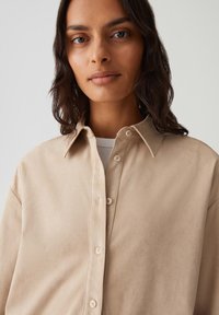 Vrouw met een gemiddelde huidtint en donker golvend haar, die een beige knopenhemd over een witte geribbelde top draagt, op een effen lichte achtergrond.