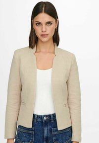 Beige herringbone-bleiseri ilman kaulusta, jossa on kaksi etutaskua ja tiukka silhouette, käytetty valkoisen paidan ja tummansiniset farkut päällä.
