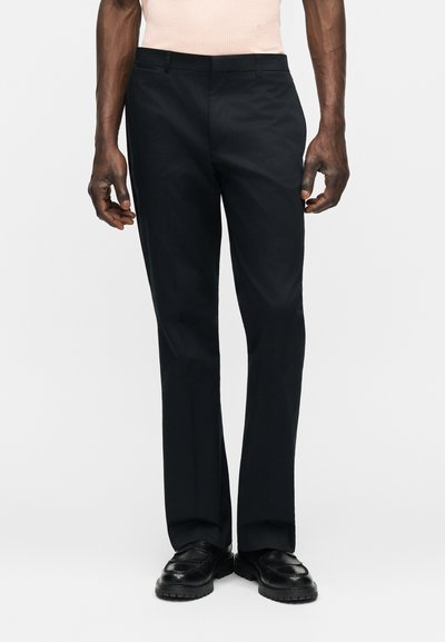 Άνδρας που φοράει εφαρμοστά μαύρα παντελόνια και μαύρα δερμάτινα μοκασίνια, στέκεται μπροστά σε απλό λευκό φόντο.