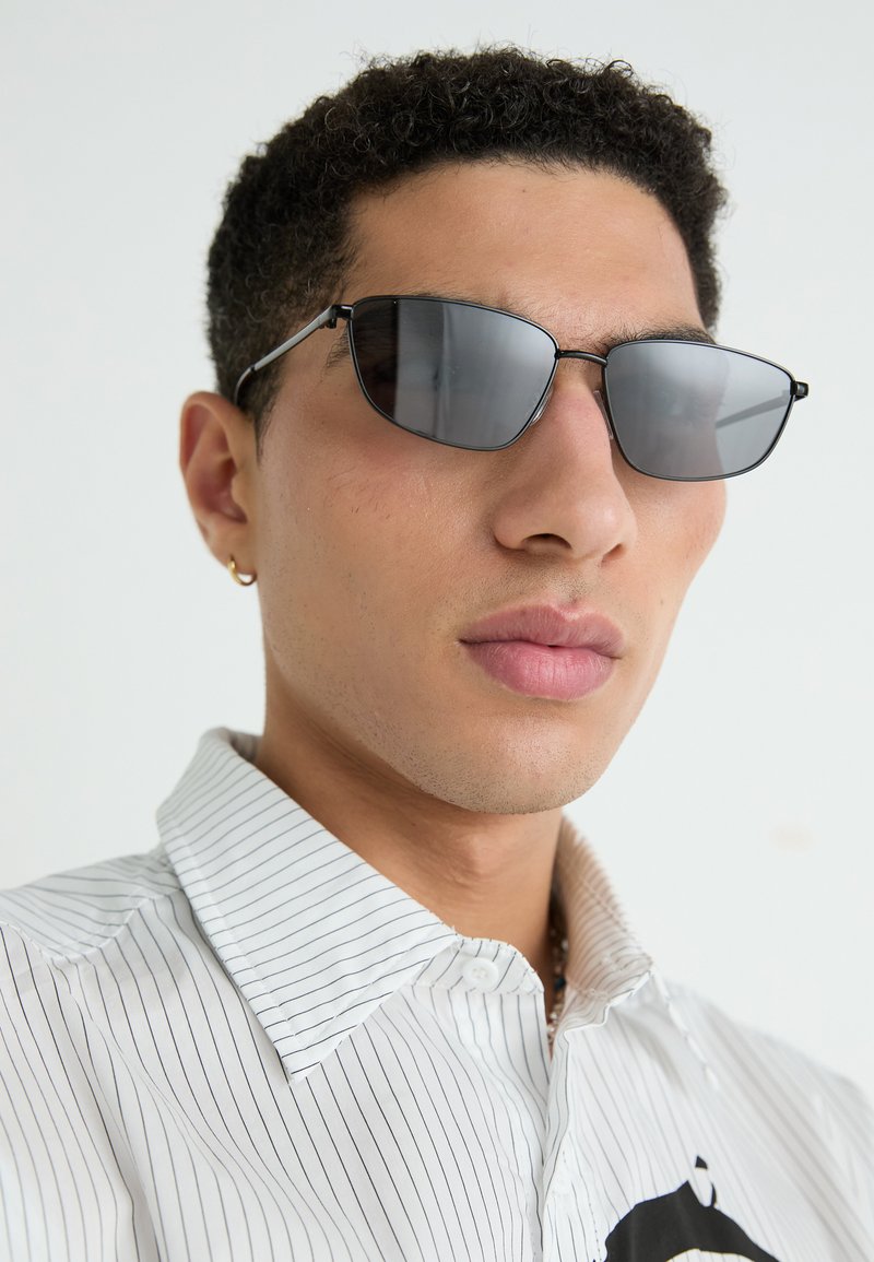 Schwarze rechteckige Sonnenbrille mit dünnem metallischen Rahmen, dunklen Gläsern und sleekem Design. Getragen mit einem gestreiften Hemd.