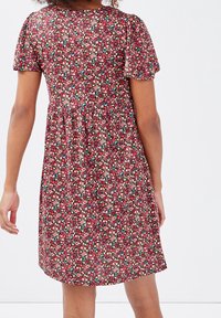 Femme portant une robe courte à manches courtes, longueur genou, avec un motif floral rouge, vue de dos sur un fond blanc uni.