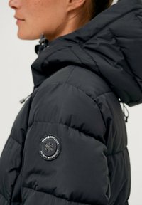 Zwarte pufferjack met een glanzende afwerking, voorzien van een capuchon, gevoerd textuur en een rond logo-oppatch op de linkerschouder.