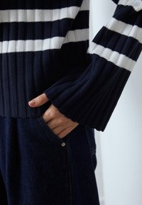 Le pull côtelé bleu marine présente des rayures horizontales blanches. La main du modèle repose dans une poche en denim foncé, mettant en valeur un simple bouton en métal.