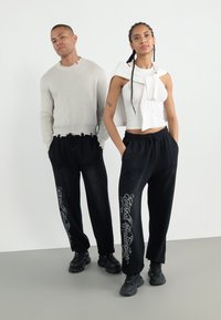 Two models stand side by side. The male model wears a light gray sweater with frayed edges and black pants, while the female model wears a white tank top with a bow and black pants featuring white lettering.