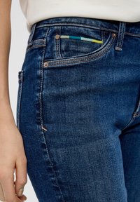 Jeans en denim bleu foncé à coupe près du corps, avec une petite poche mettant en valeur un détail tissé multicolore. Couture de contraste légère le long des coutures.