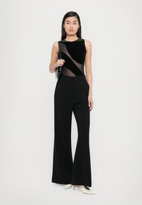 Black sleeveless top with sheer paneling, paired with high-waisted black flared trousers and beige pointed-toe heels. Minimalist black bag.