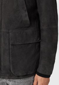 Gros plan sur une veste en daim noir montrant une grande poche frontale, une manche longue avec poignet, et une partie de la main et un jean gris.