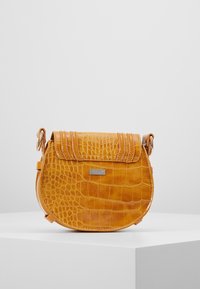 Borsa a tracolla in ecopelle coccodrillo color cammello con pattina, dettagli di cucitura, piastra con logo in metallo e tracolla regolabile su superficie bianca.