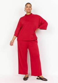 Červený pletený dvojdielny outfit so voľným vysokým golierom, širokými rukávmi a širokými nohavicami. Dopĺňajú ho čierne nazúvacie topánky.