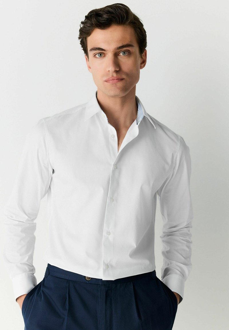 Cămașă albă cu dungi, cu guler tip button-down, mâneci lungi și detalii albastre în contrast în interiorul gulerului. Pur tată cu pantaloni bleumarin.