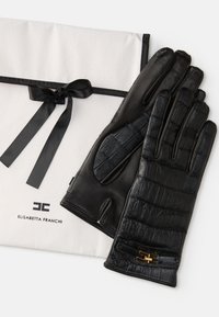 Luvas de couro negro com textura de crocodilo. Apresentam um detalhe em metal dourado e estão embaladas numa bolsa de tecido branca com uma fita preta.