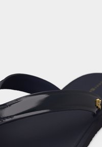 Σαγιονάρα σε ναυτικό μπλε με γυαλιστερό λουράκι και μικρό χρυσό λογότυπο της μάρκας στο πλάι, σε κοντινή λήψη πάνω σε λευκό φόντο.