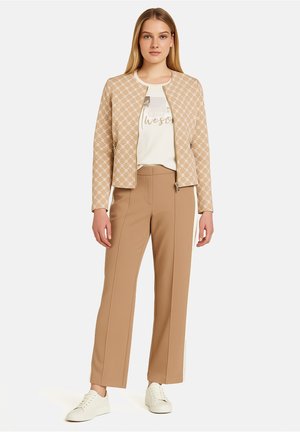 Veste beige à motifs avec fermeture éclair, associée à un t-shirt blanc avec texte, et un pantalon beige droit. Baskets blanches.