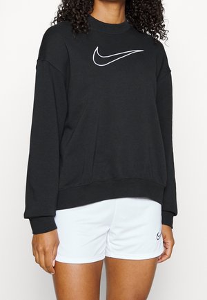 Osoba nosząca czarną, długą bluzę Nike z białym logo Swoosh oraz białe spodenki Nike z logo Swoosh na lewej nodze.