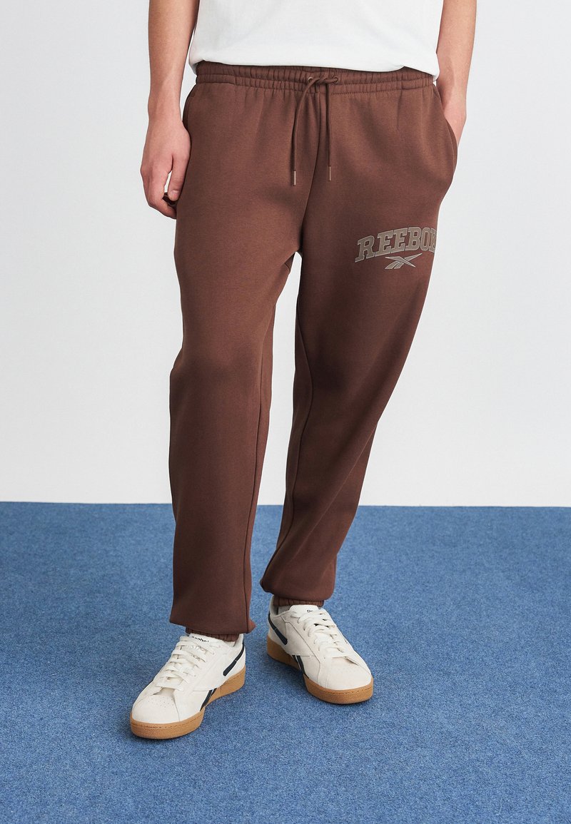Persona con pantalones de chándal marrones de Reebok y zapatillas blancas de pie sobre una alfombra azul, con las manos en los bolsillos, frente a un fondo de pared blanca.