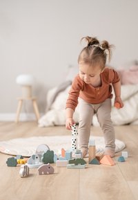 Houten blokken in verschillende vormen en kleuren, waaronder groene bomen, roze en blauwe dieren, en stukjes met stippen op een lichte vloer.