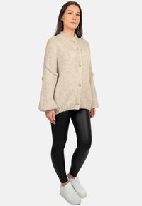 Cardigan beige lavorato a maglia spesso con colletto alto, bottoni grandi e maniche svasate, abbinato a leggings neri e sneakers bianche.