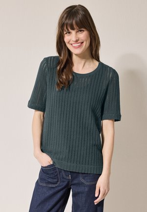 Dunkelgrüner gestrickter Kurzarmpullover mit strukturiertem, offenen Muster, rundem Ausschnitt und lockerer Passform. Kombiniert mit dunklen Denim-Jeans.