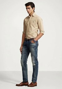 Jaunas vyras stovi su viena ranka kišenėje, vilkėdamas smėlio spalvos ilgomis rankovėmis marškinį, nusidėvėjusius mėlynus džinsus, rudą diržą, laikrodį ir rudus loaferius.