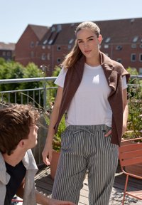 Brązowa kamizelka bez rękawów na białej koszulce, zestawiona z szarymi spodniami w paski. Model stoi na balkonie z zielonymi roślinami i budynkami w tle.