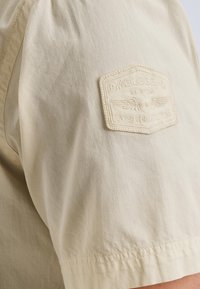 Chemise beige à manches courtes avec une texture côtelée et un badge brodé "FM LEGEND" sur la manche gauche. Coutures visibles au niveau du poignet.